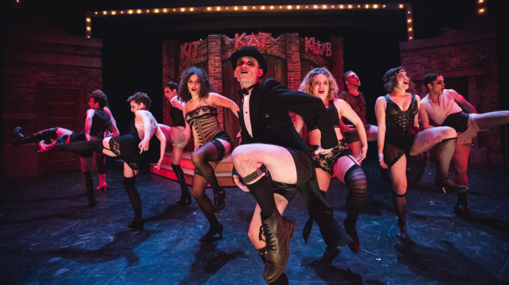 John Drea as the Emcee in Cabaret. He strikes a post onstage with an ensemble of dancers behind him.